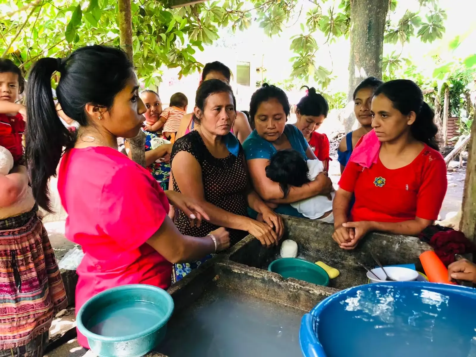 A group of women and children gathered around a water basin as a facilitator demonstrates clean water practices in an outdoor community setting.